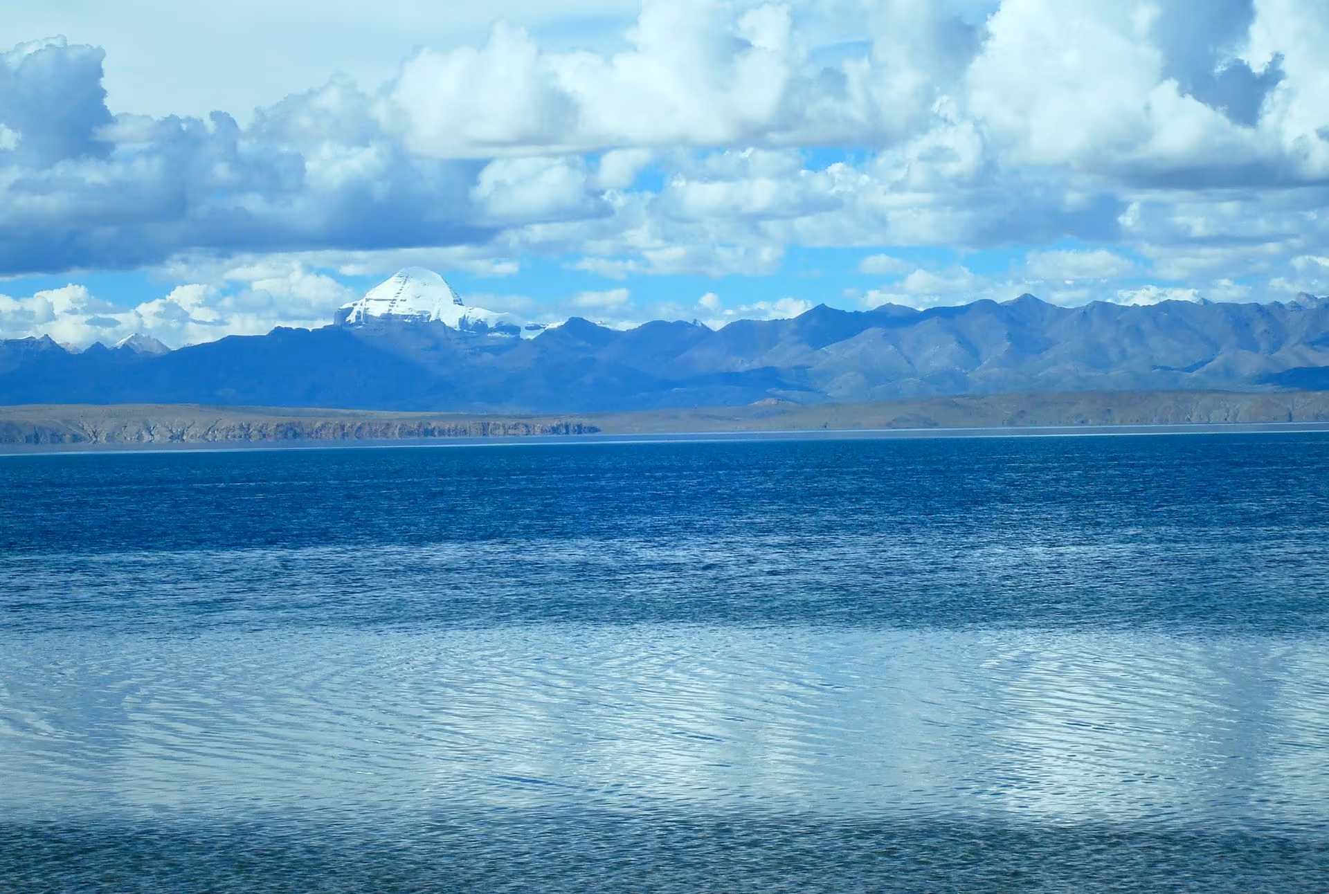 Mount Kailash small and white on the horizon beyond the blue water of Lake Manasarovar