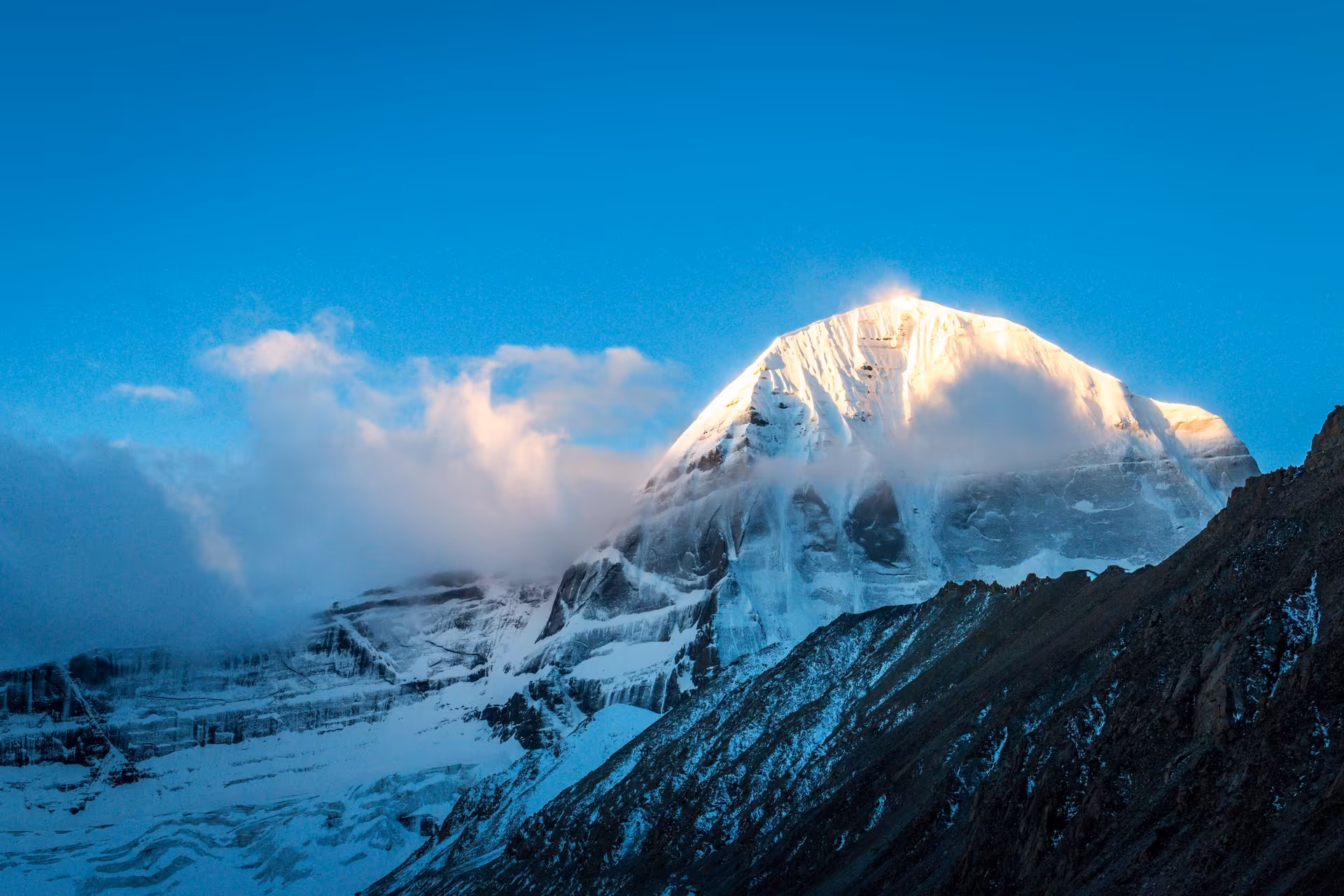 The snow-covered face of Mount Kailash catching low golden light above a bank of cloud